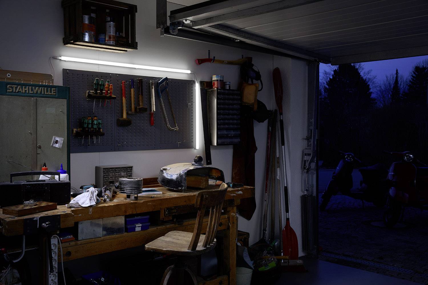 Un atelier éclairé la nuit. Un établi avec des outils, une chaise en bois, des étagères et un garage ouvert donnant sur l'obscurité.