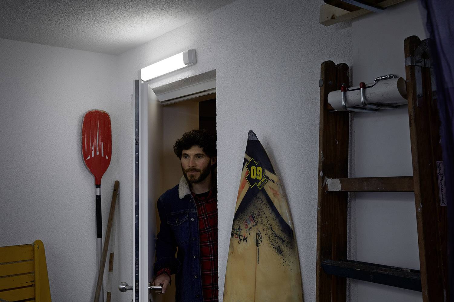 Un homme entre par une porte dans une pièce contenant des planches de surf, des pagaies et une échelle. L'éclairage est allumé.