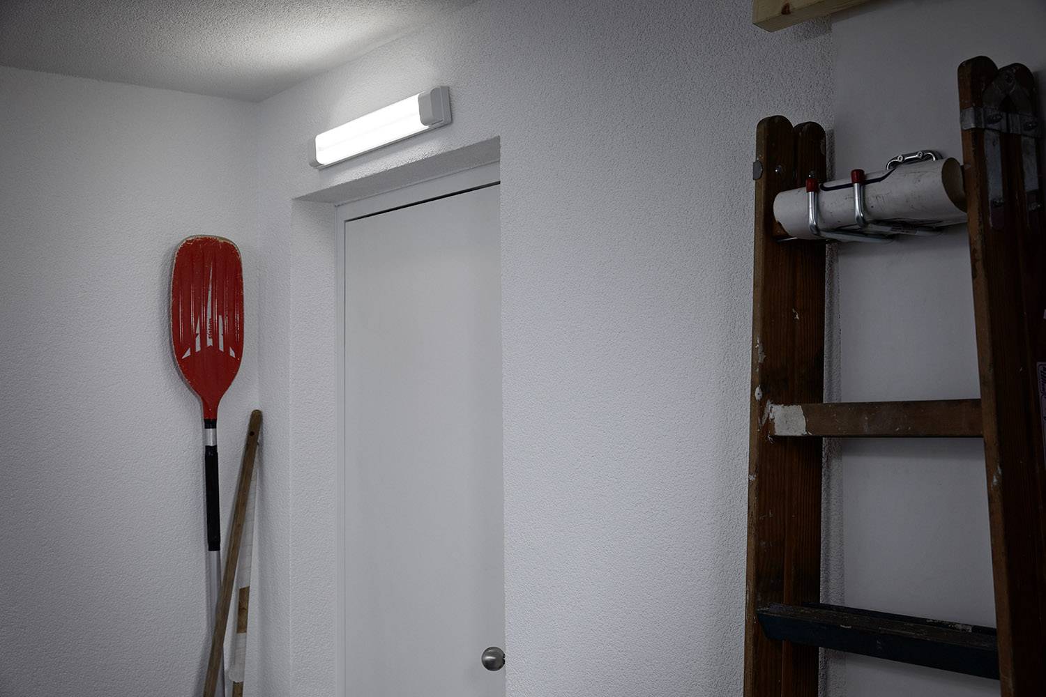 Un cadre de porte fermé dans une pièce avec un mur blanc, une pagaie rouge accrochée au mur et une échelle en bois à droite. En haut, une lumière brille.