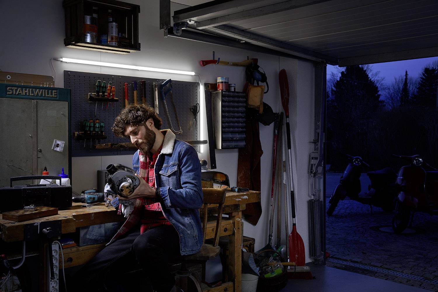 Un homme travaille avec concentration sur un morceau de métal dans un atelier. Le garage est bien organisé avec des outils accrochés au mur. À l'extérieur, il fait déjà noir.