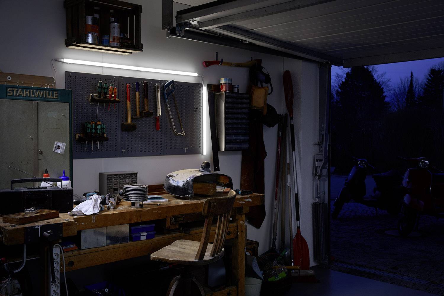 Atelier dans un garage ouvert la nuit, avec une table de travail éclairée, des outils suspendus à un support mural, et une moto à l'extérieur.