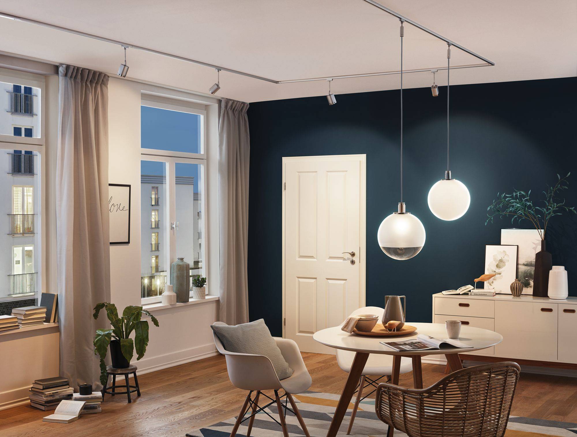 Un salon moderne avec un sol en bois, une grande fenêtre, une table et des chaises. Des lampes sont accrochées au mur, et à travers la fenêtre, on aperçoit des bâtiments illuminés.