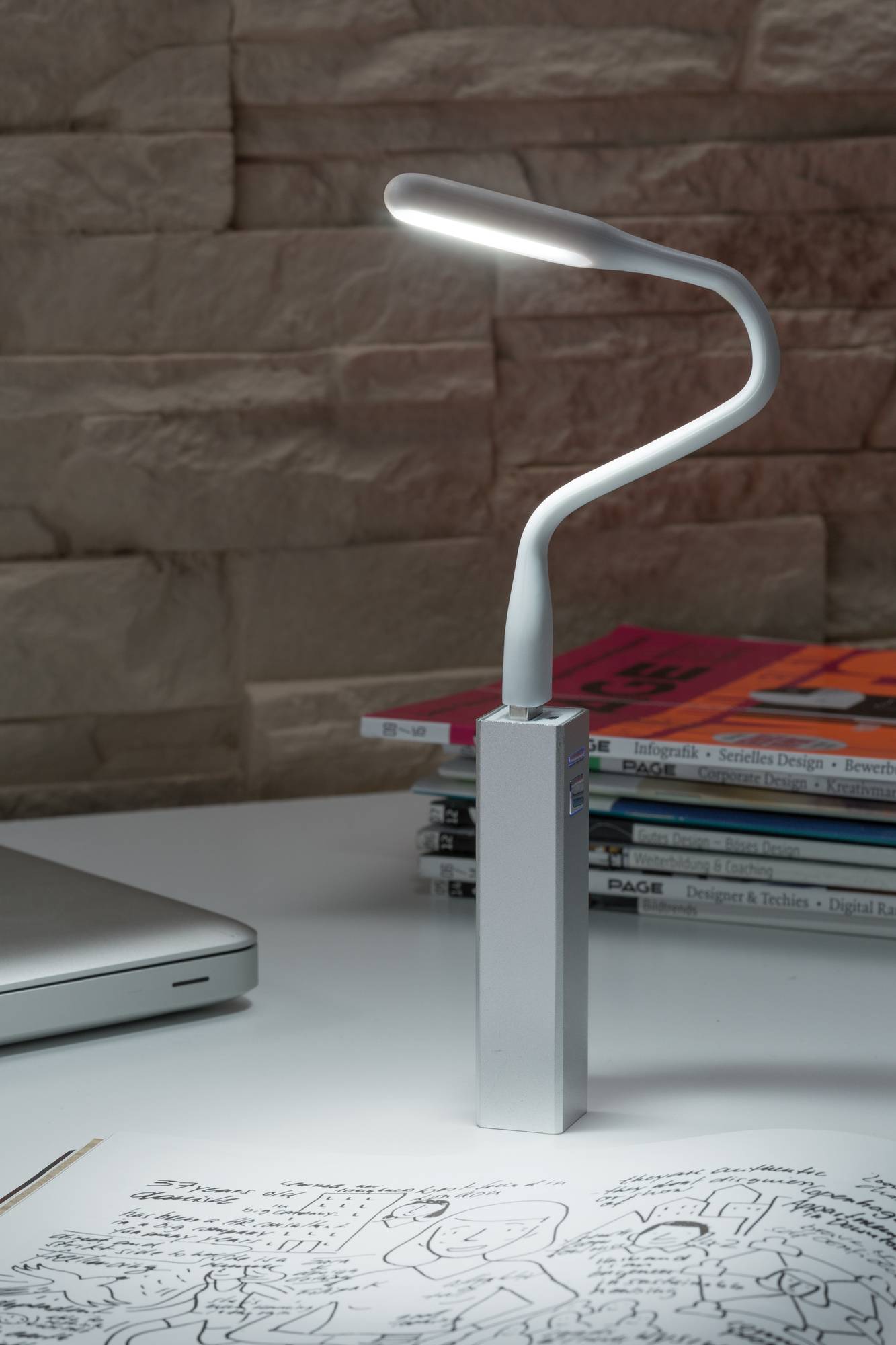 Bureau avec une lampe LED blanche moderne éclairant un carnet de notes. À côté, un ordinateur portable et des piles de magazines sont posés.