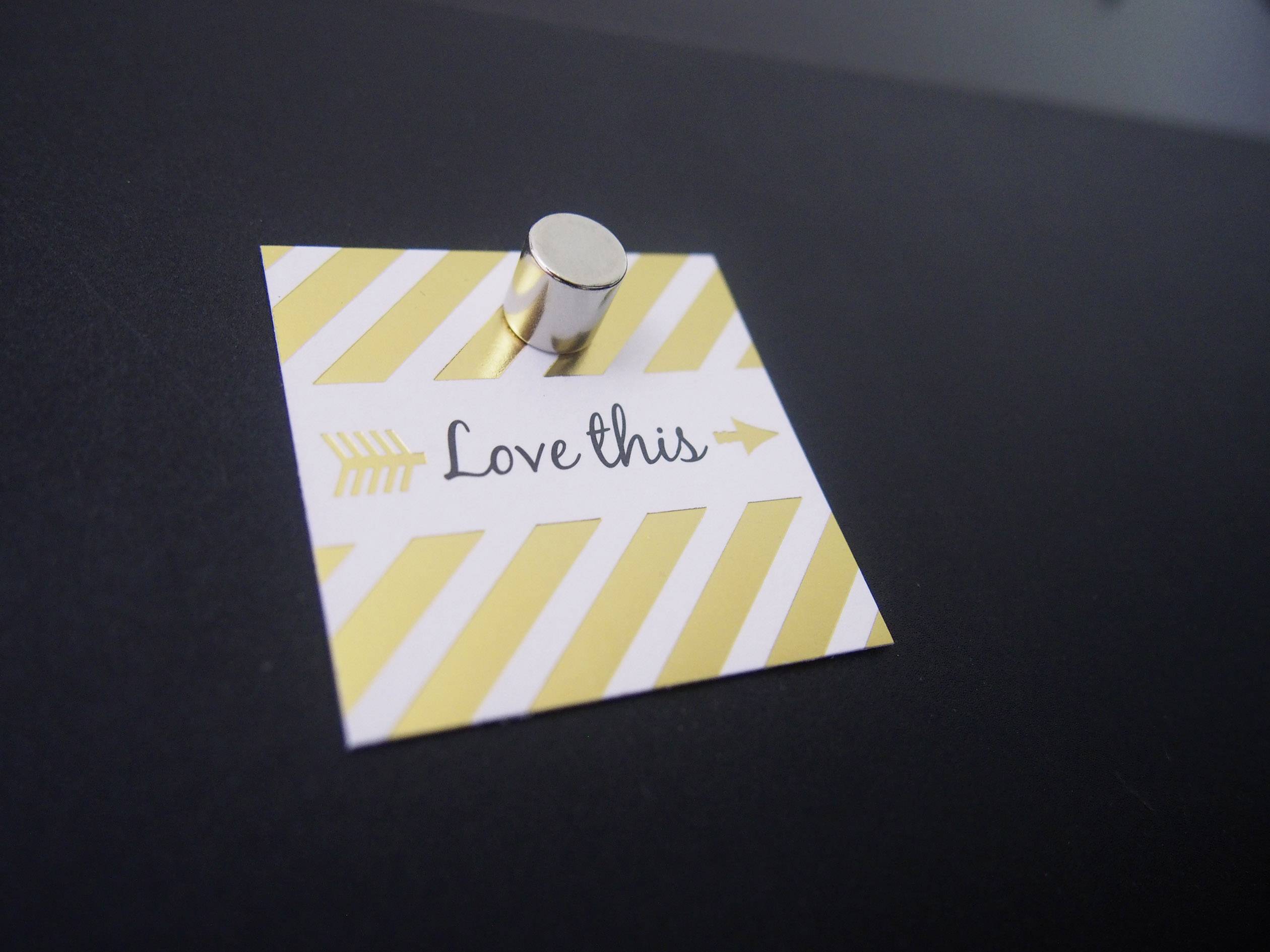 Petit post-it sur fond noir, présentant des rayures jaunes diagonales avec l'inscription « Love this ». Fixé par un aimant argenté.