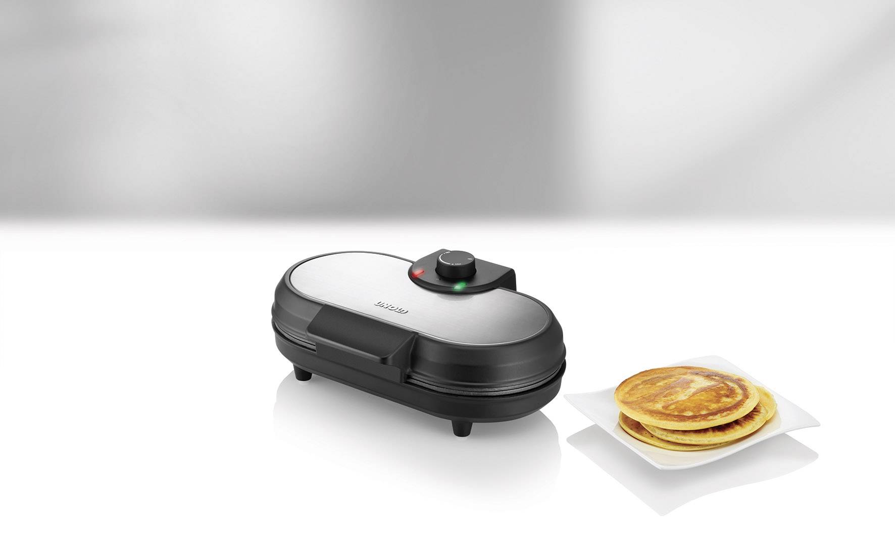 Un gaufrier argenté avec témoin lumineux est posé sur un fond blanc. À côté se trouve une assiette garnie de pancakes dorés et croustillants.