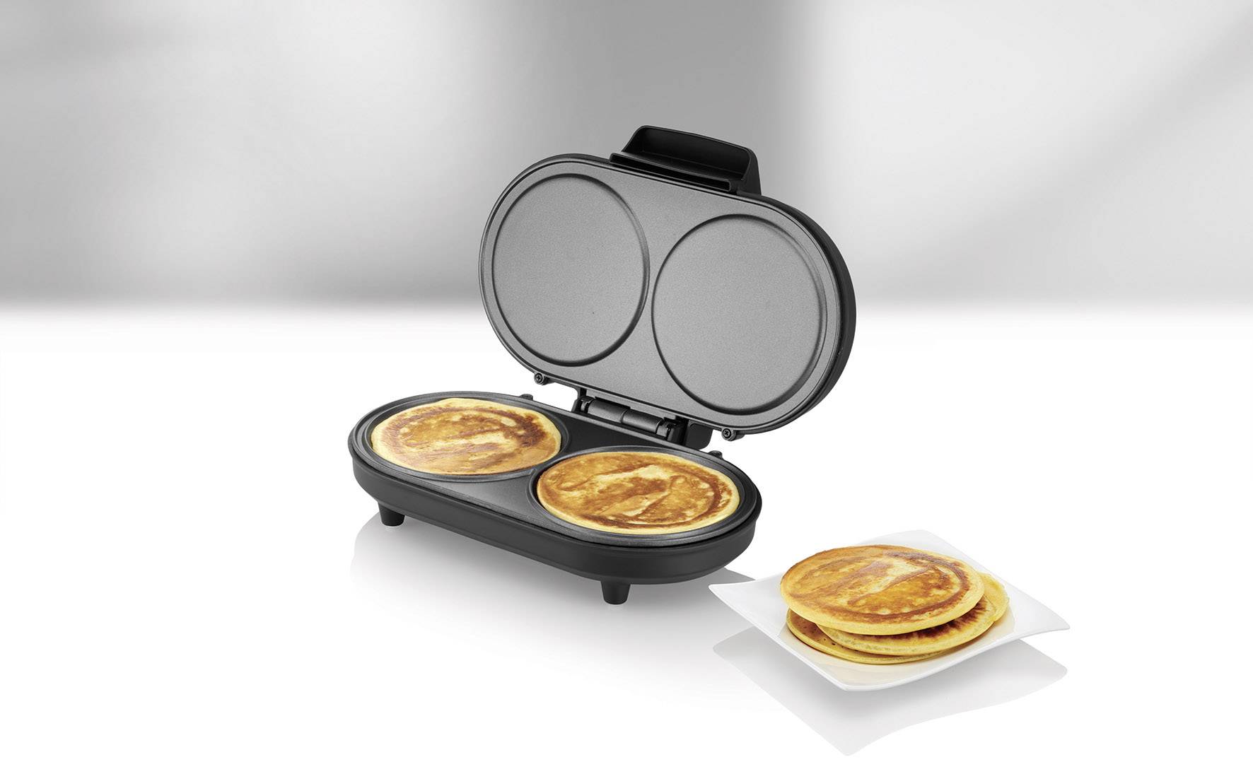 Un gaufrier avec deux gaufres rondes, à côté d'une assiette empilée de gaufres. Arrière-plan gris-blanc neutre.