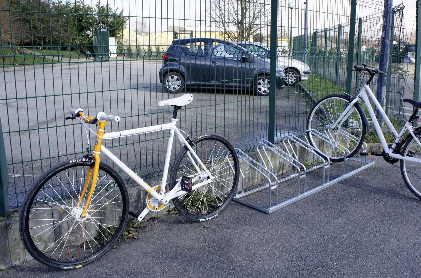 Deux vélos, l'un contre une clôture et l'autre sur un support, devant un parking avec une voiture garée en arrière-plan.