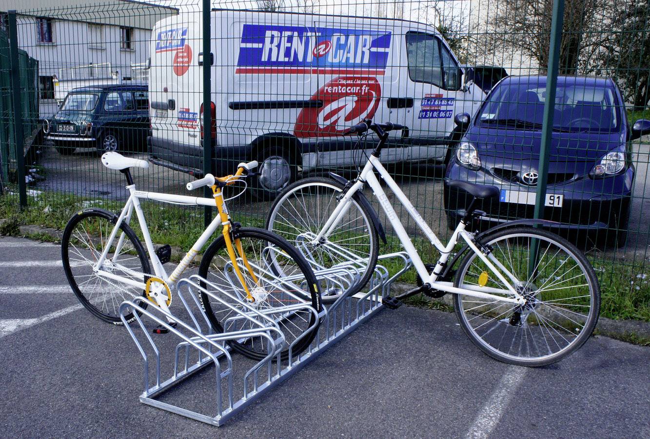 Deux vélos sont garés dans un support à vélos. À l'arrière-plan se trouve un fourgon blanc portant l'inscription 'RENT A CAR'.