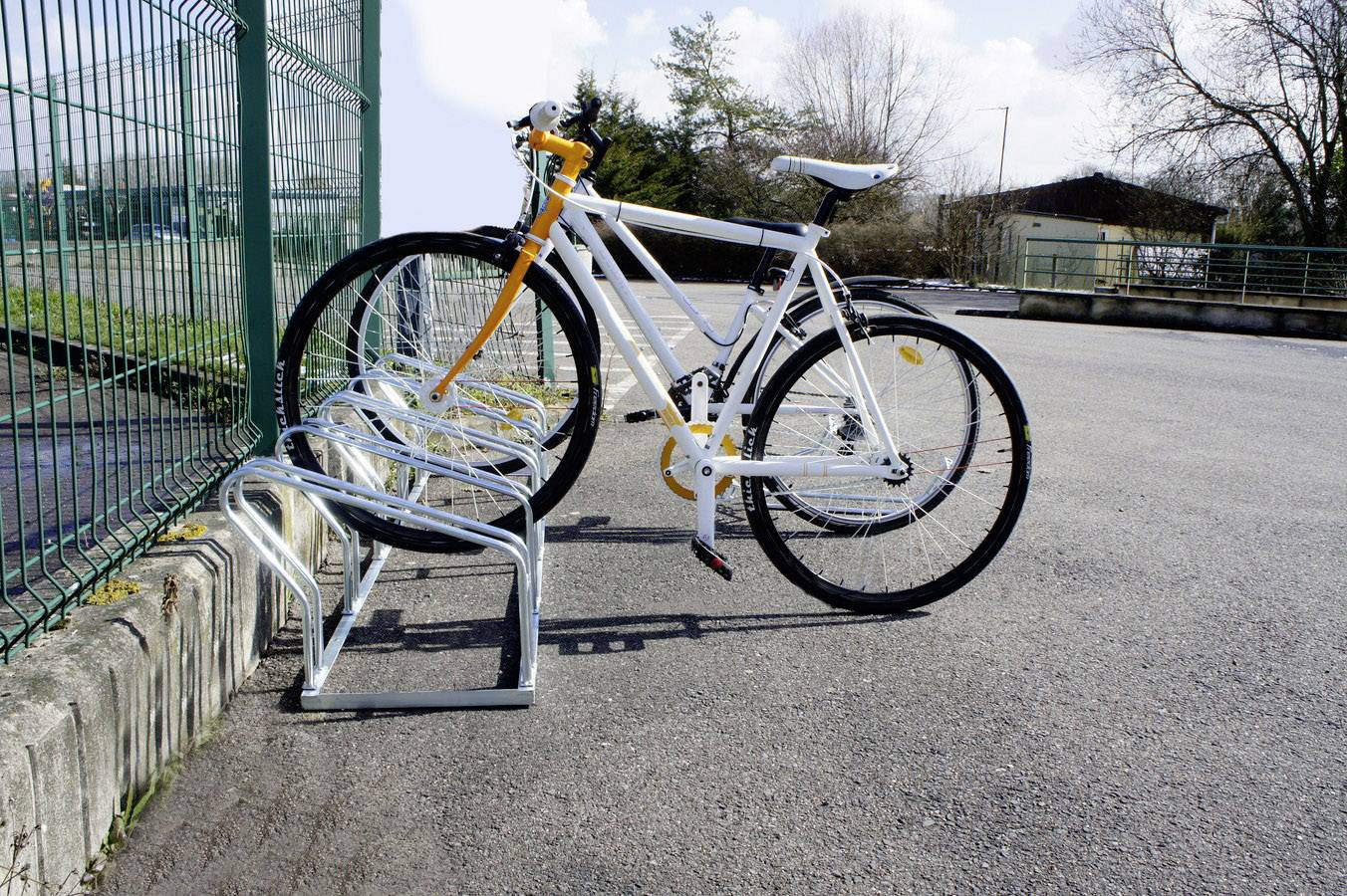 Un vélo blanc est stationné dans un support à vélos sur un parking vide. Des arbres et une clôture sont visibles en arrière-plan.