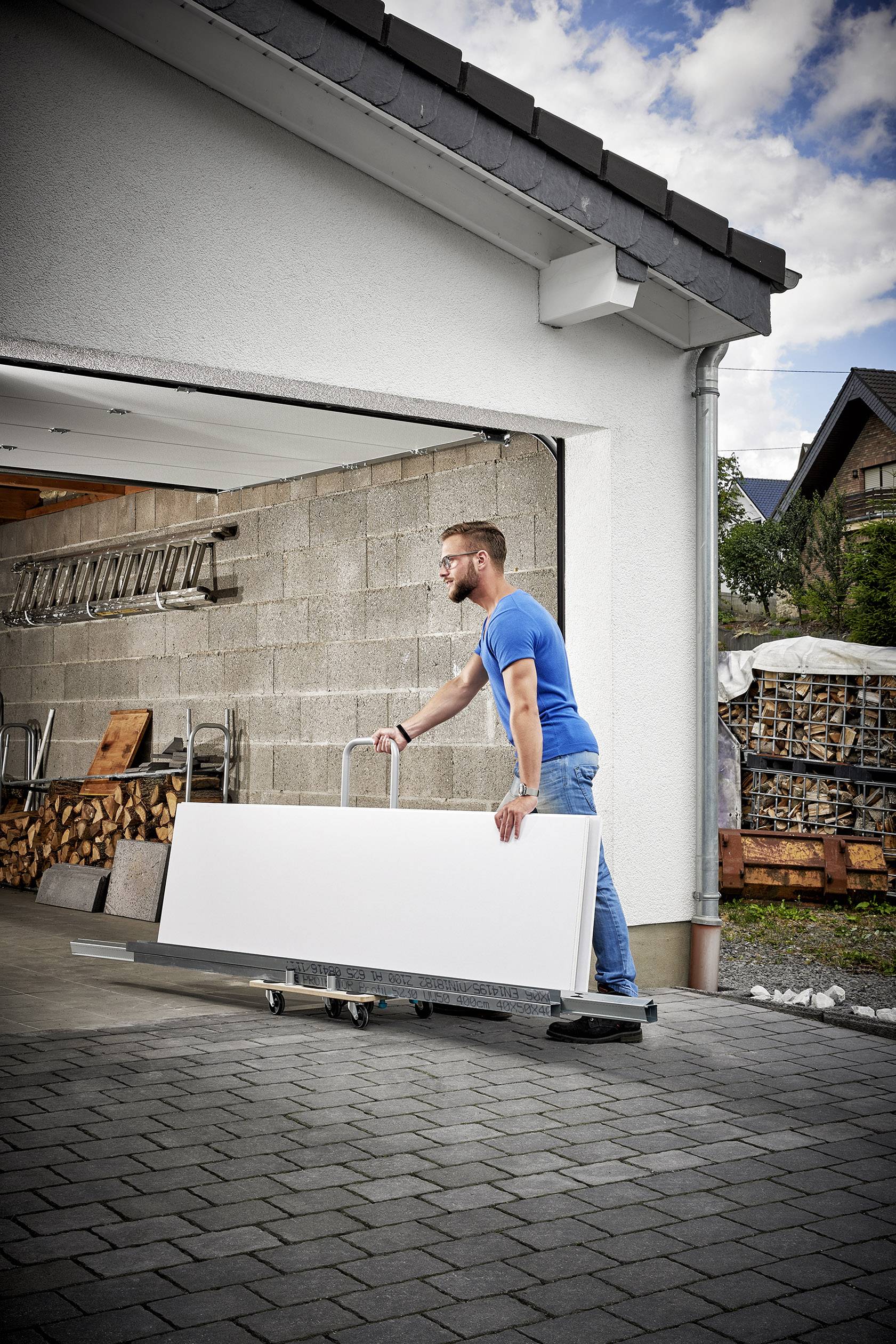 Un homme portant un t-shirt bleu déplace un grand panneau blanc sur un chariot roulant vers un garage. Des piles de bois et une échelle sont visibles en arrière-plan.