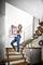 Une femme descend un grand carton dans un escalier, souriante. Environnement moderne avec des escaliers en bois et un arrangement de plantes en arrière-plan.