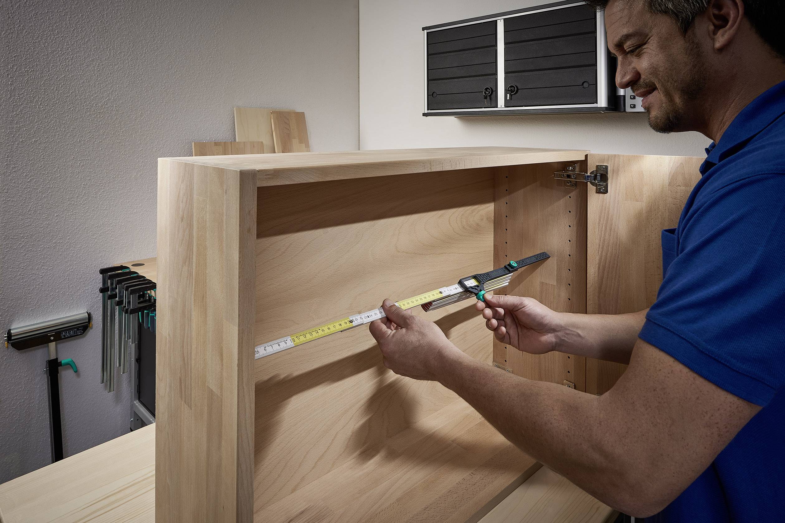 Un homme mesure la largeur intérieure d'un placard en bois à l'aide d'un mètre ruban dans un atelier. Des outils et des planches de bois sont visibles en arrière-plan.