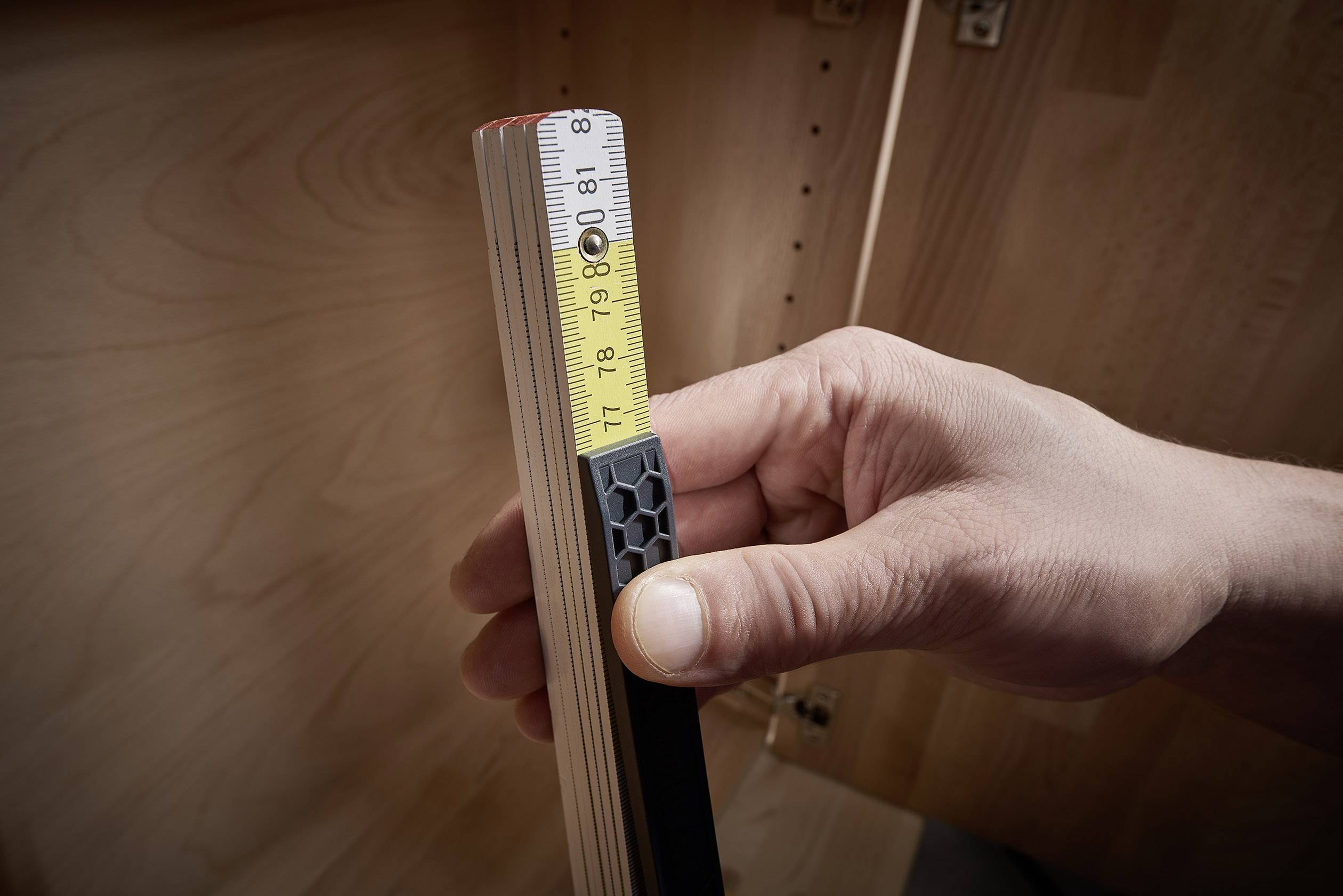 Une main tient un mètre pliant en bois, affichant une longueur d'environ 9 pouces. Des meubles en bois sont visibles en arrière-plan.