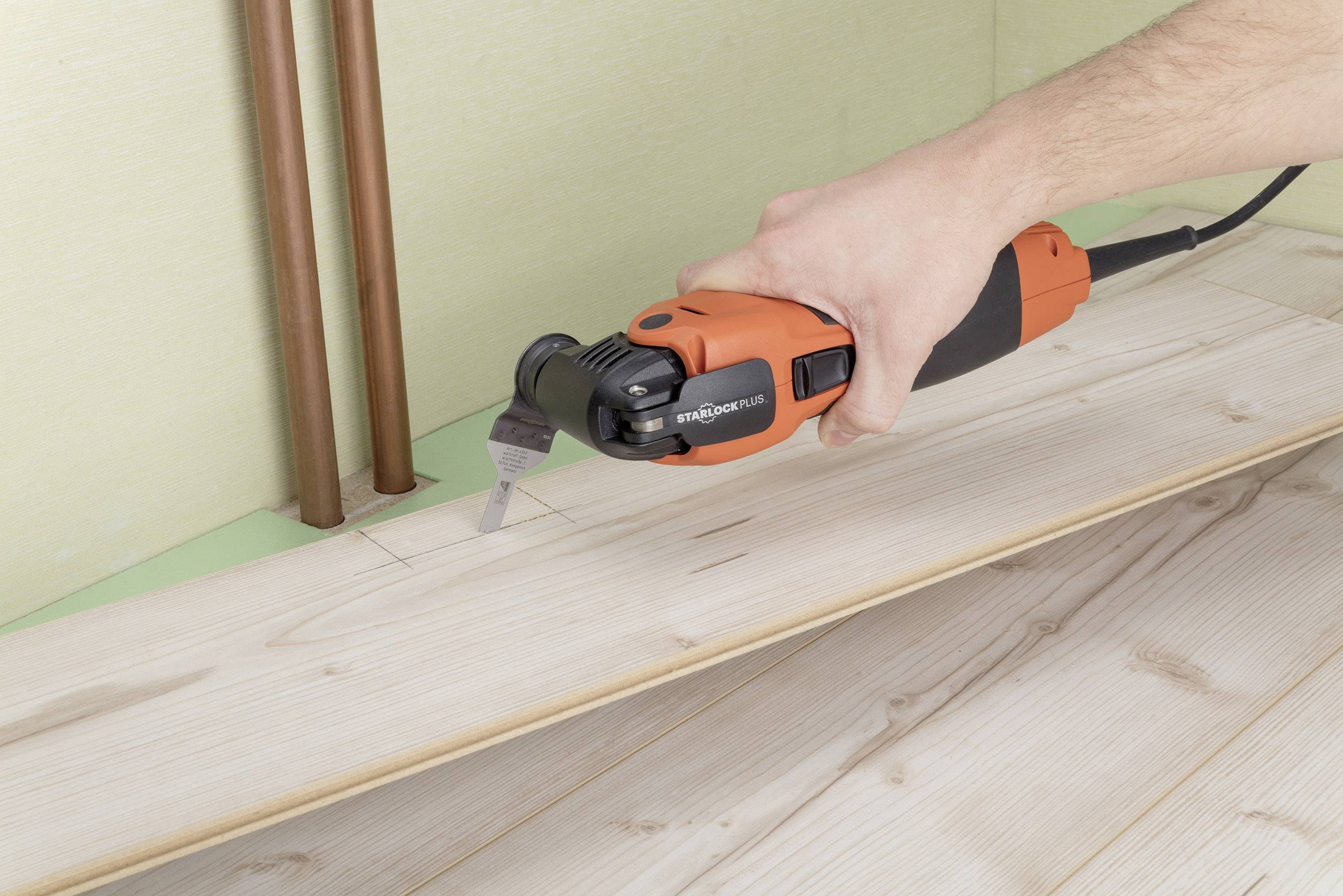 Une personne découpe une encoche dans une planche de parquet à l'aide d'un outil multifonction oscillant près de tuyaux.