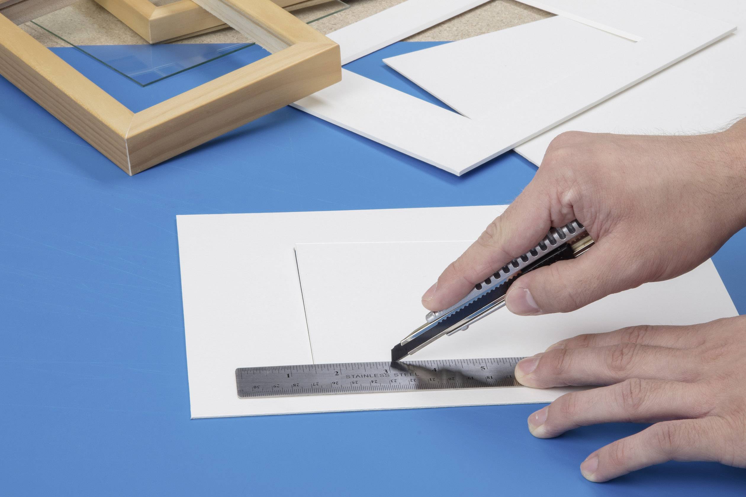 Une main découpe un rectangle avec un cutter dans du papier blanc sur un support bleu, à côté d'une règle et d'un cadre.