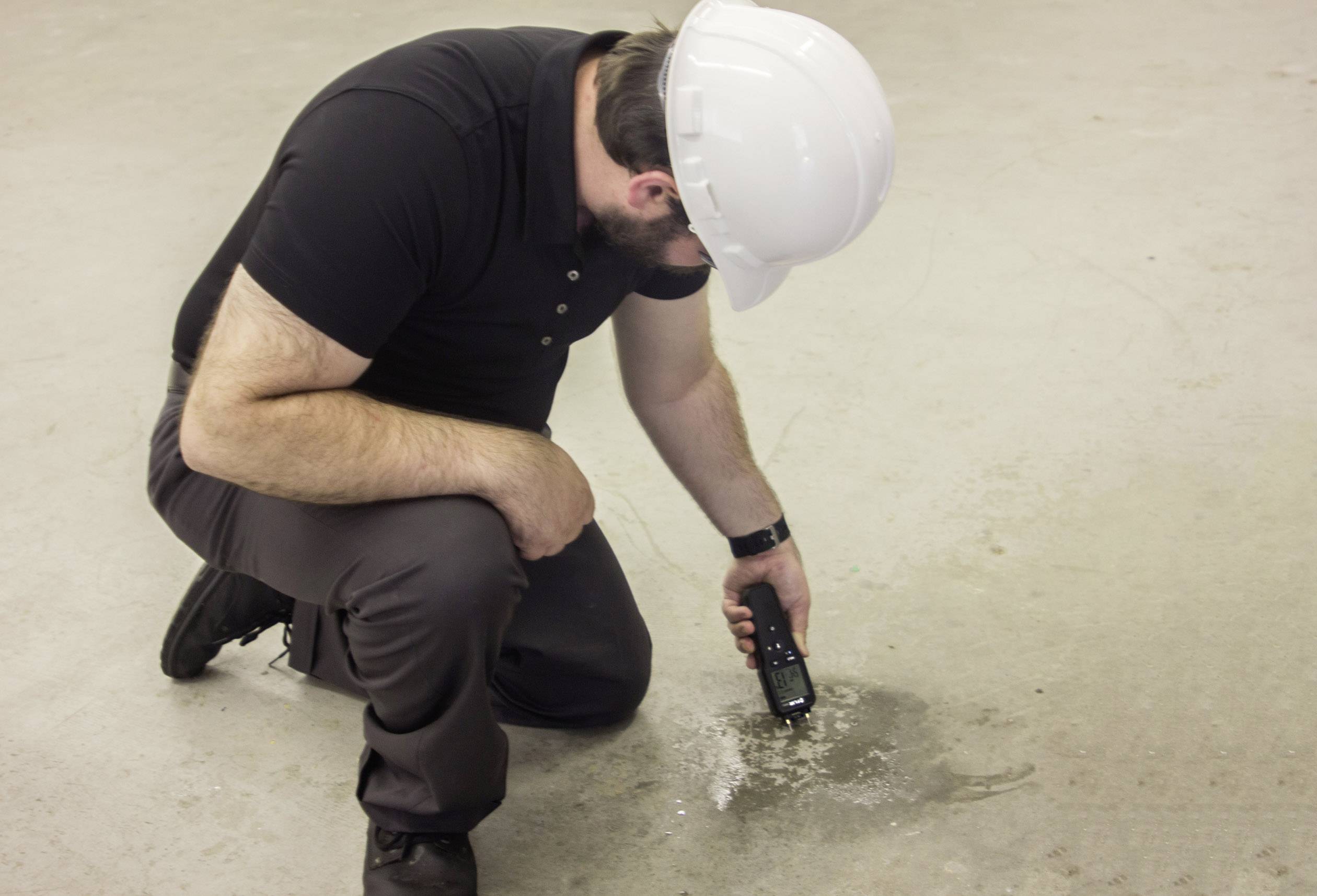 Une personne portant un casque de chantier est agenouillée au sol et mesure l'humidité d'une flaque avec un appareil portable.