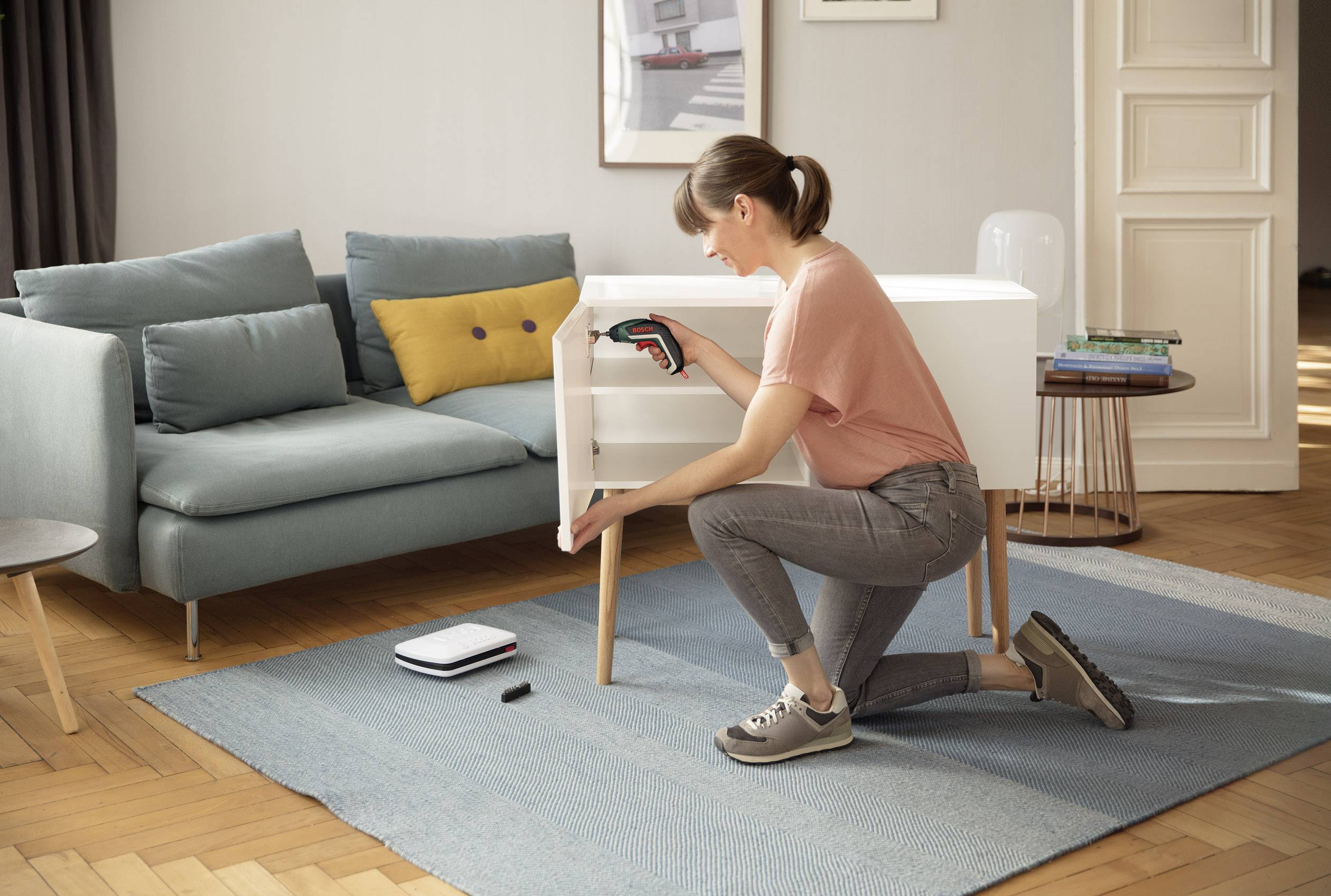 Une femme est agenouillée sur un tapis bleu dans le salon et assemble un meuble avec un tournevis sans fil. Un canapé est visible en arrière-plan.