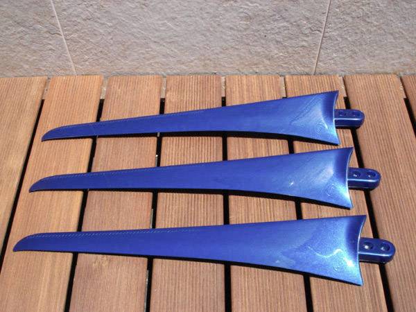 Trois ailerons de surf bleus, aérodynamiques, sont disposés parallèlement sur un sol en bois ; ils sont brillants et légèrement incurvés.