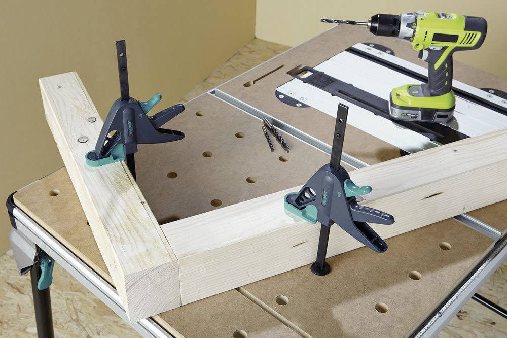 Sur une table, un cadre en bois est fixé avec deux serre-joints. Une perceuse et des vis sont posées à côté. Situation de poste de travail.