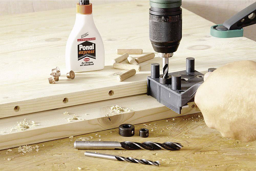 Une main utilise un gabarit de perçage et une perceuse pour faire des trous dans du bois. Des chevilles en bois et de la colle à bois sont posées sur la table.