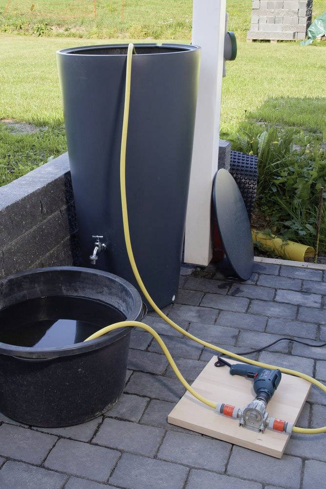 Un grand réservoir d'eau gris à l'extérieur avec un tuyau jaune qui dirige l'eau vers un seau noir. Une pompe est posée à côté.