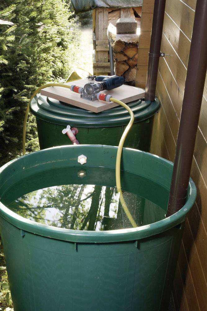 Deux citernes vertes de récupération d'eau de pluie placées à côté d'une maison, reliées par des tuyaux et une pompe pour utiliser l'eau de pluie collectée.