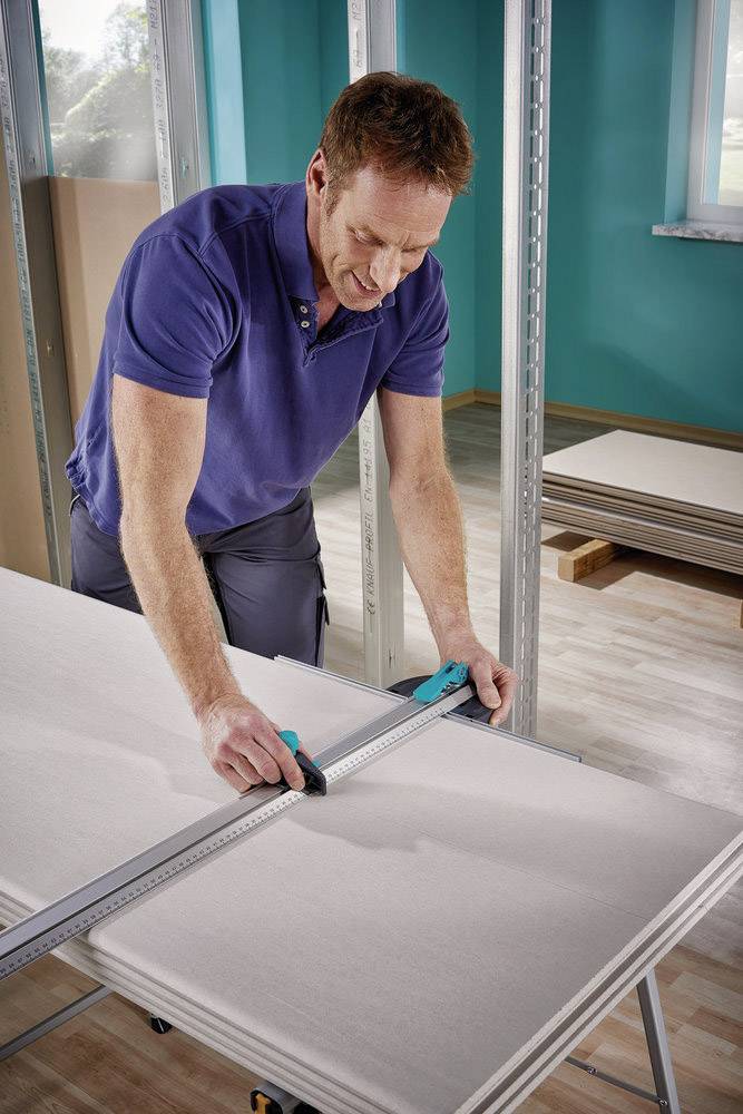Un homme mesure avec une règle et un outil de coupe sur une plaque de plâtre dans une pièce en construction ou en rénovation.