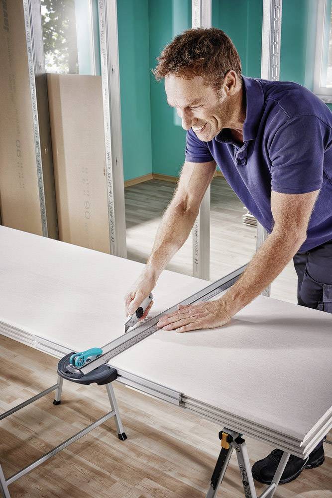 Un homme portant une chemise bleue coupe une plaque de plâtre avec un cutter sur une table dans une pièce en construction.