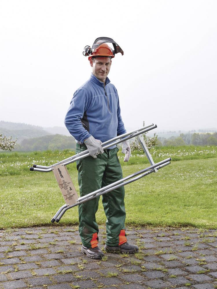 Une personne en tenue de protection se tient dehors sur un chemin pavé, tenant un support métallique dans sa main, avec de l'herbe verte en arrière-plan.
