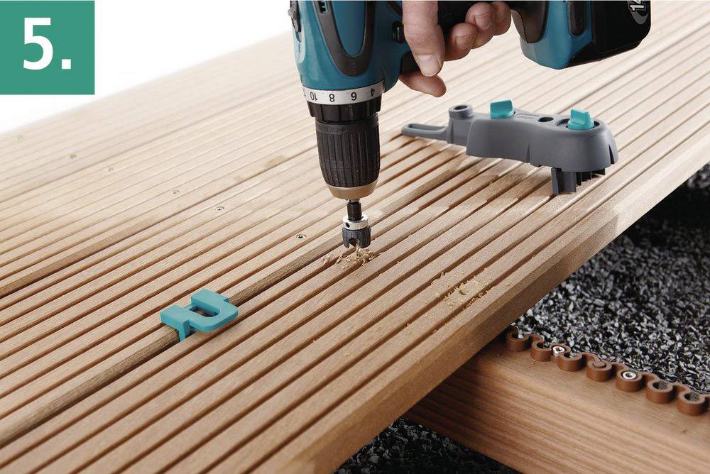 Une personne utilise une visseuse sans fil pour percer des trous dans une terrasse en bois. Des cales et un outil de fixation sont posés sur la terrasse.