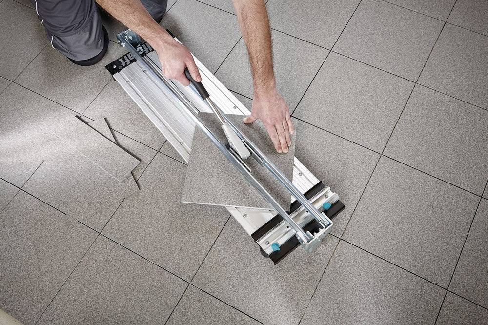 Une personne coupe un carreau avec un coupe-carrelage. Les carreaux sont disposés en motif quadrillé sur le sol.