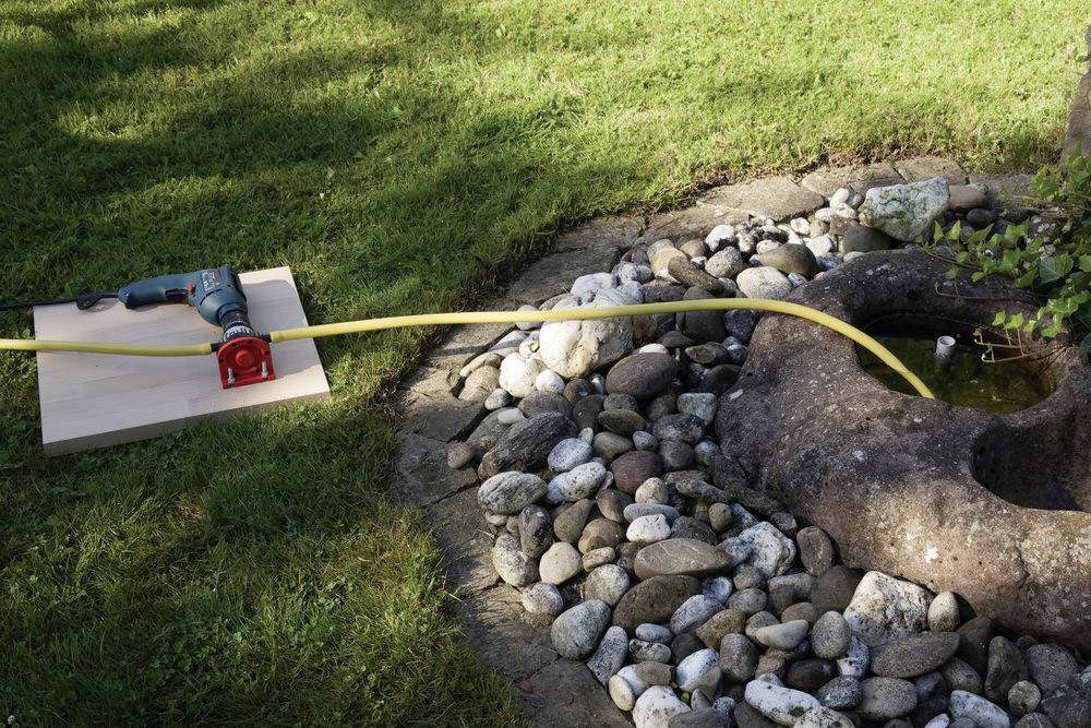 Perceuse avec un tuyau pompant de l'eau dans un puits en pierre, entouré de galets et d'herbe.