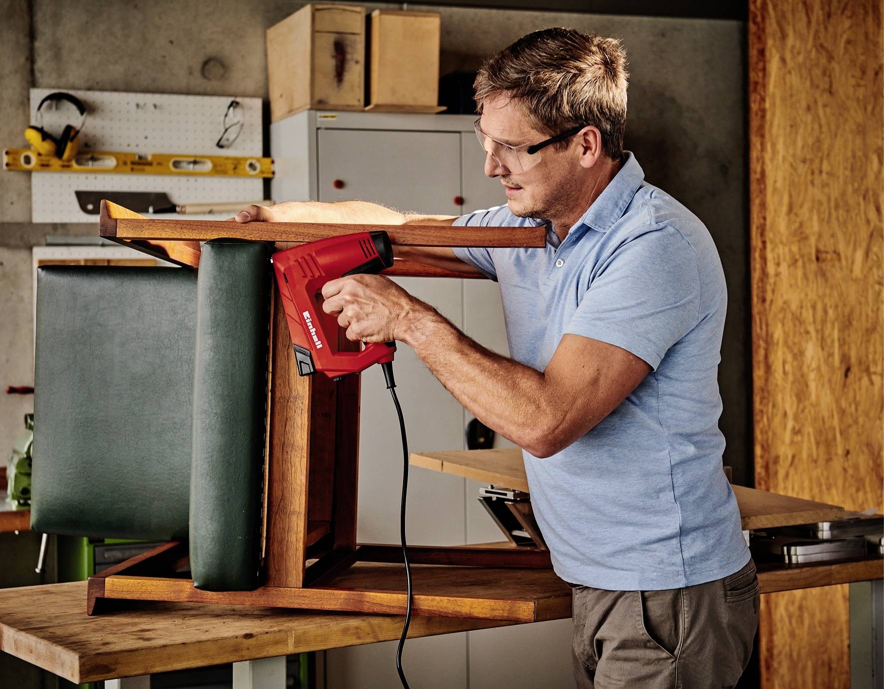 Un homme dans un atelier agrafait un nouveau coussin sur une chaise retournée. Il porte des lunettes de protection et utilise un pistolet à agrafes rouge.