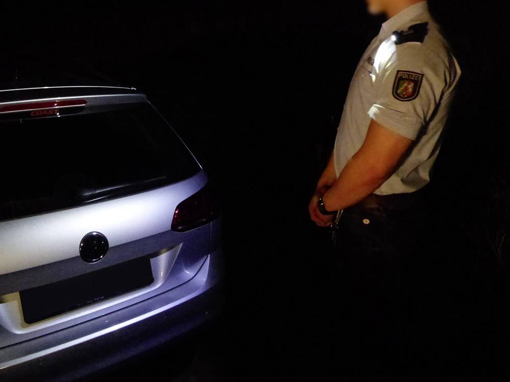 Une personne en uniforme se tient à côté d'une voiture garée dans un environnement sombre. La scène est éclairée par une lampe de poche.