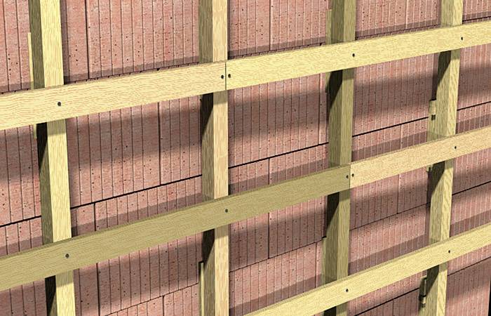 Des lattes de bois sont fixées horizontalement sur un mur de briques, probablement dans le cadre d'une construction ou d'une rénovation.