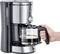 Une main tient une carafe en verre remplie de café fraîchement préparé, qui coule d'une cafetière en argent.