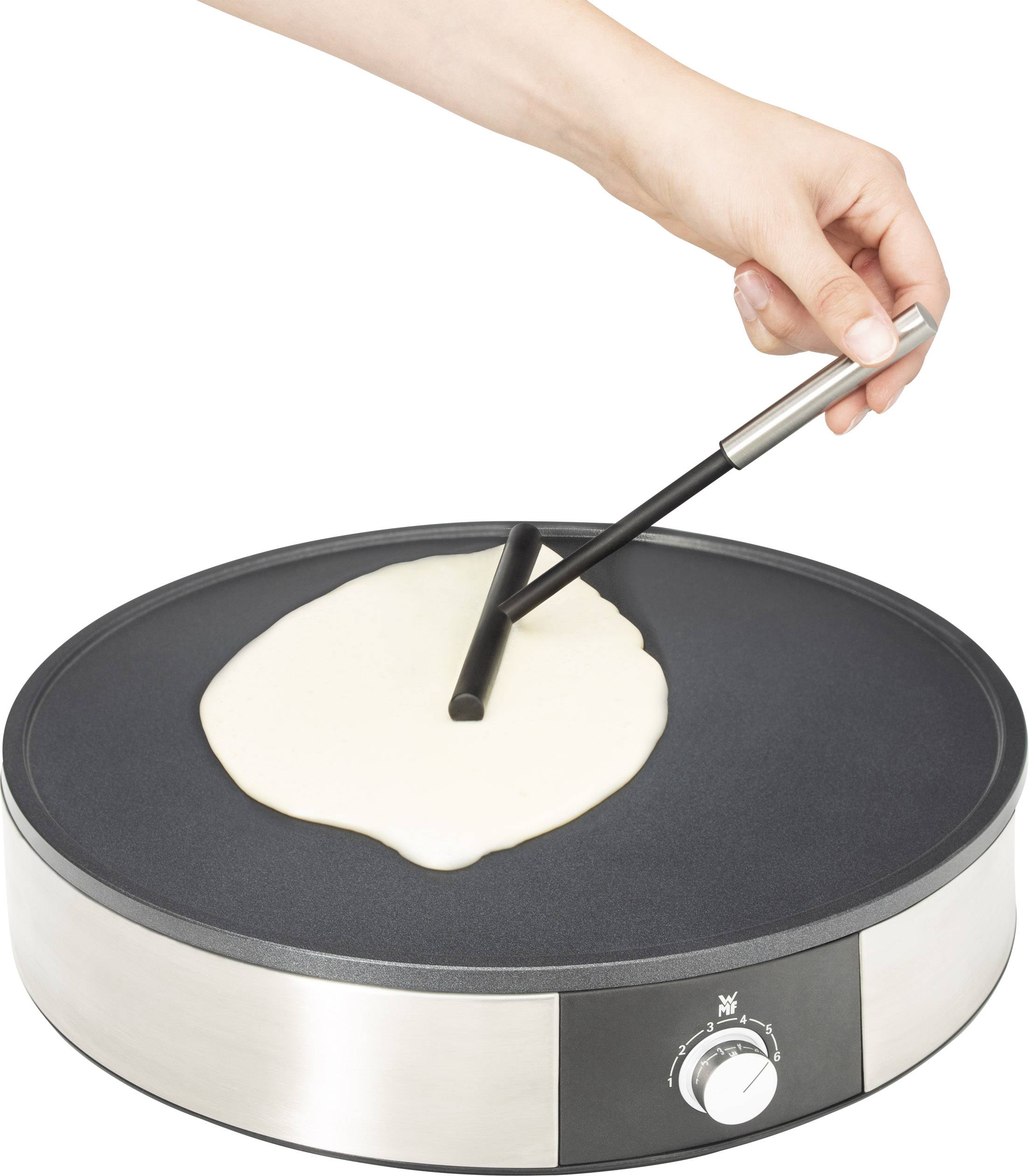 Une main répartit de la pâte liquide sur une poêle électrique ronde à crêpes à l'aide d'un répartiteur de pâte à crêpes.