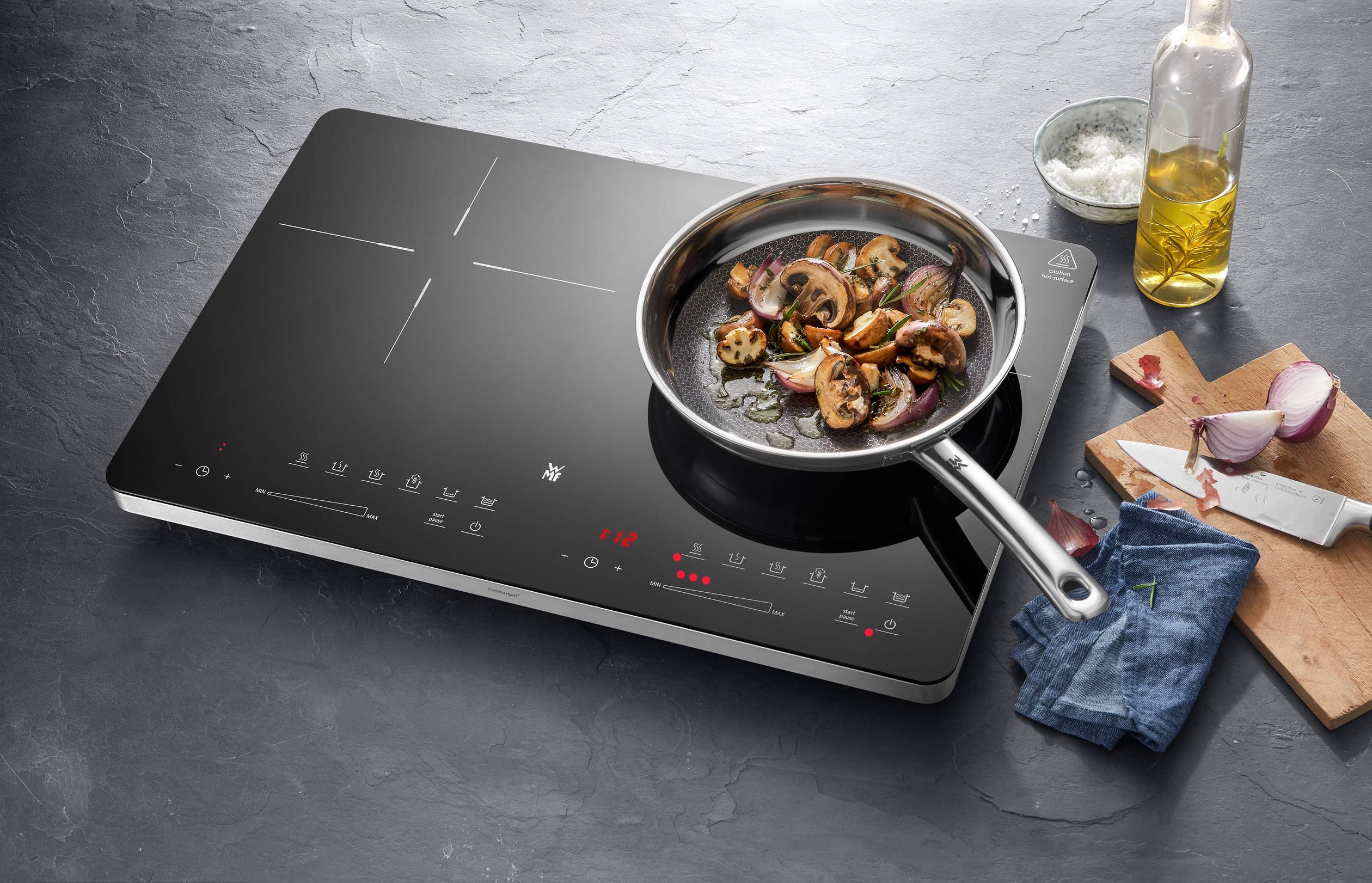 Une plaque de cuisson à induction moderne avec un plat de champignons et d'oignons. À côté, une planche à découper avec des oignons émincés.