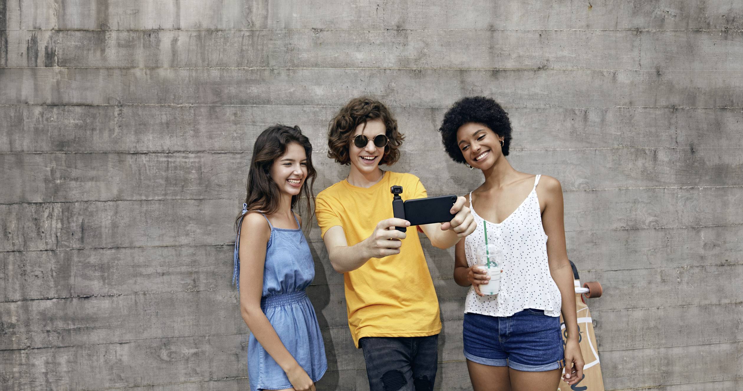 Trois jeunes adultes posent joyeusement pour un selfie devant un mur en béton. Une femme tient un longboard.