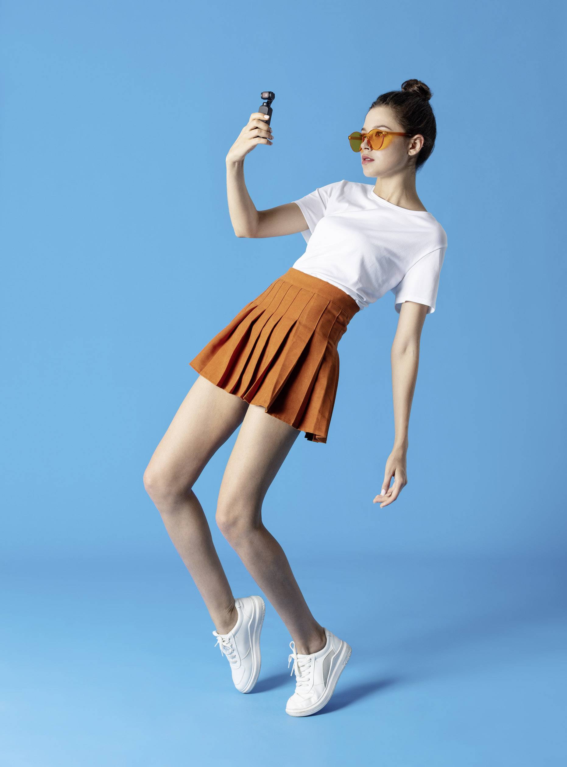 Une femme portant un t-shirt blanc et une jupe orange prend un selfie devant un fond bleu. Elle porte des lunettes de soleil et des baskets blanches.
