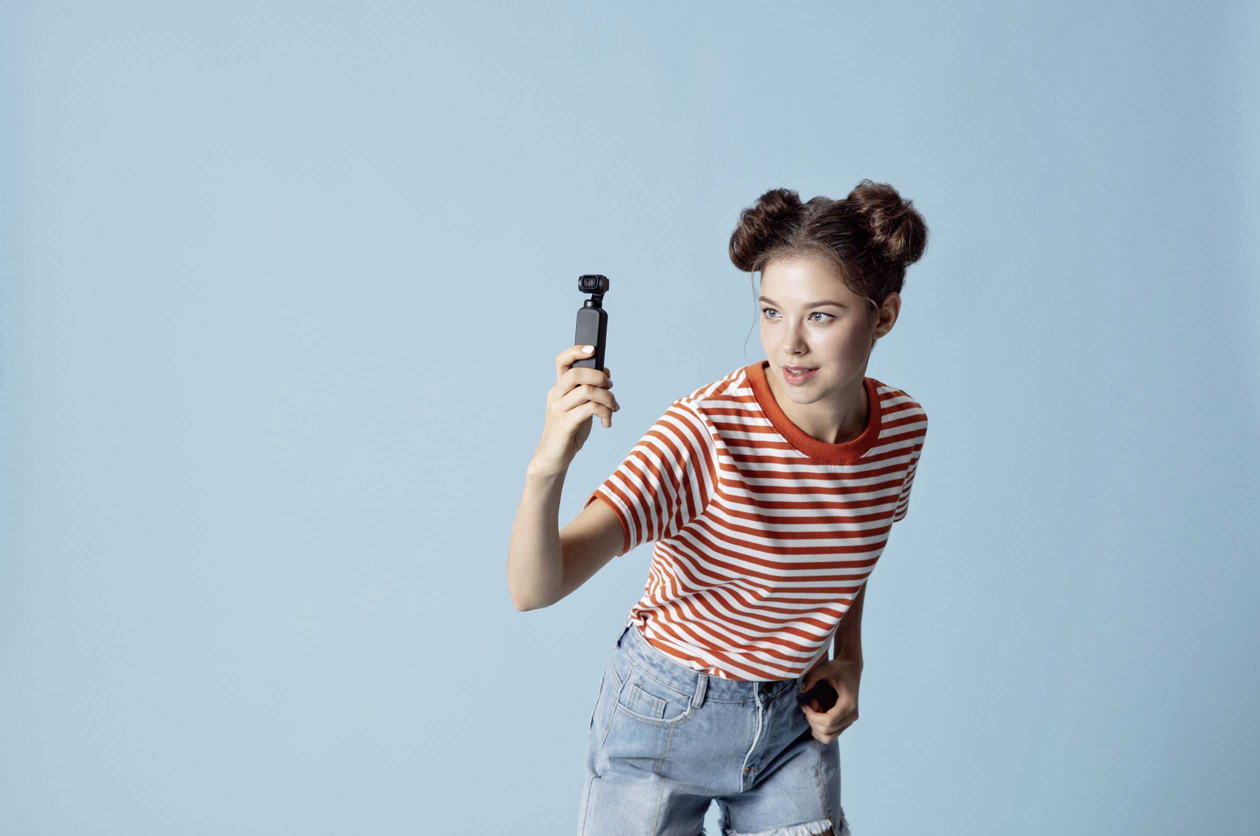 Une jeune femme portant un t-shirt rayé tient joyeusement un petit appareil photo devant un fond bleu clair.