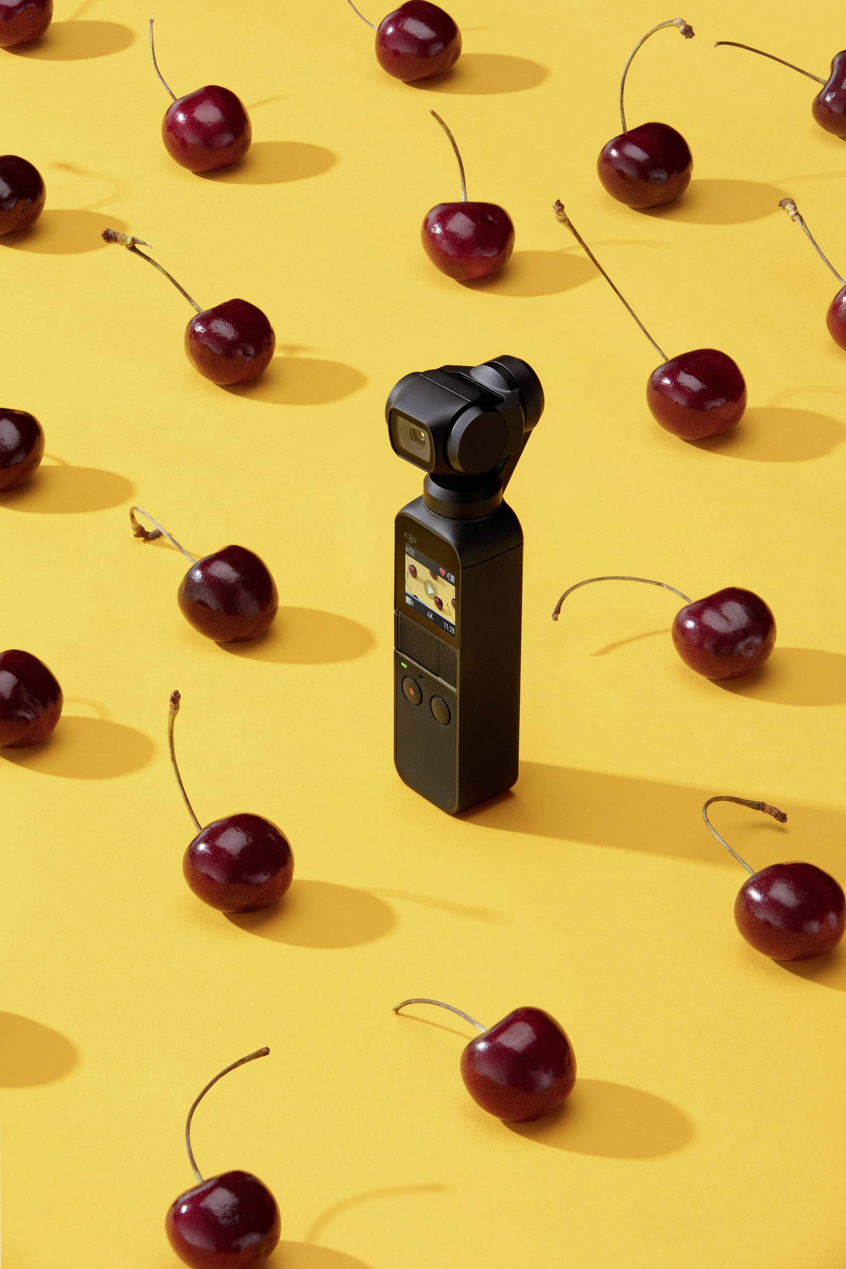 Une caméra vidéo noire est droite sur un fond jaune, entourée de cerises dispersées.