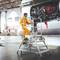 Un technicien vêtu d'une combinaison jaune travaille sur une échelle près d'un moteur d'avion ouvert dans un grand atelier lumineux.