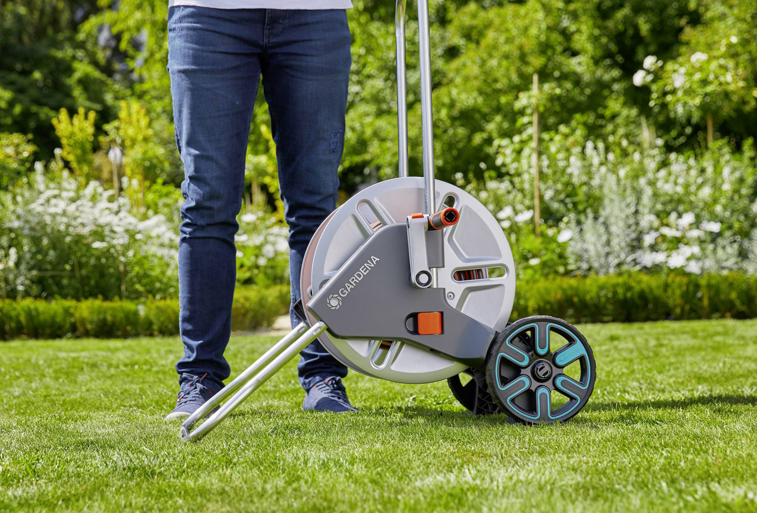 Une personne se tient sur un gazon à côté d'un dévidoir de tuyau d'arrosage. L'arrière-plan montre des plantes en fleur.