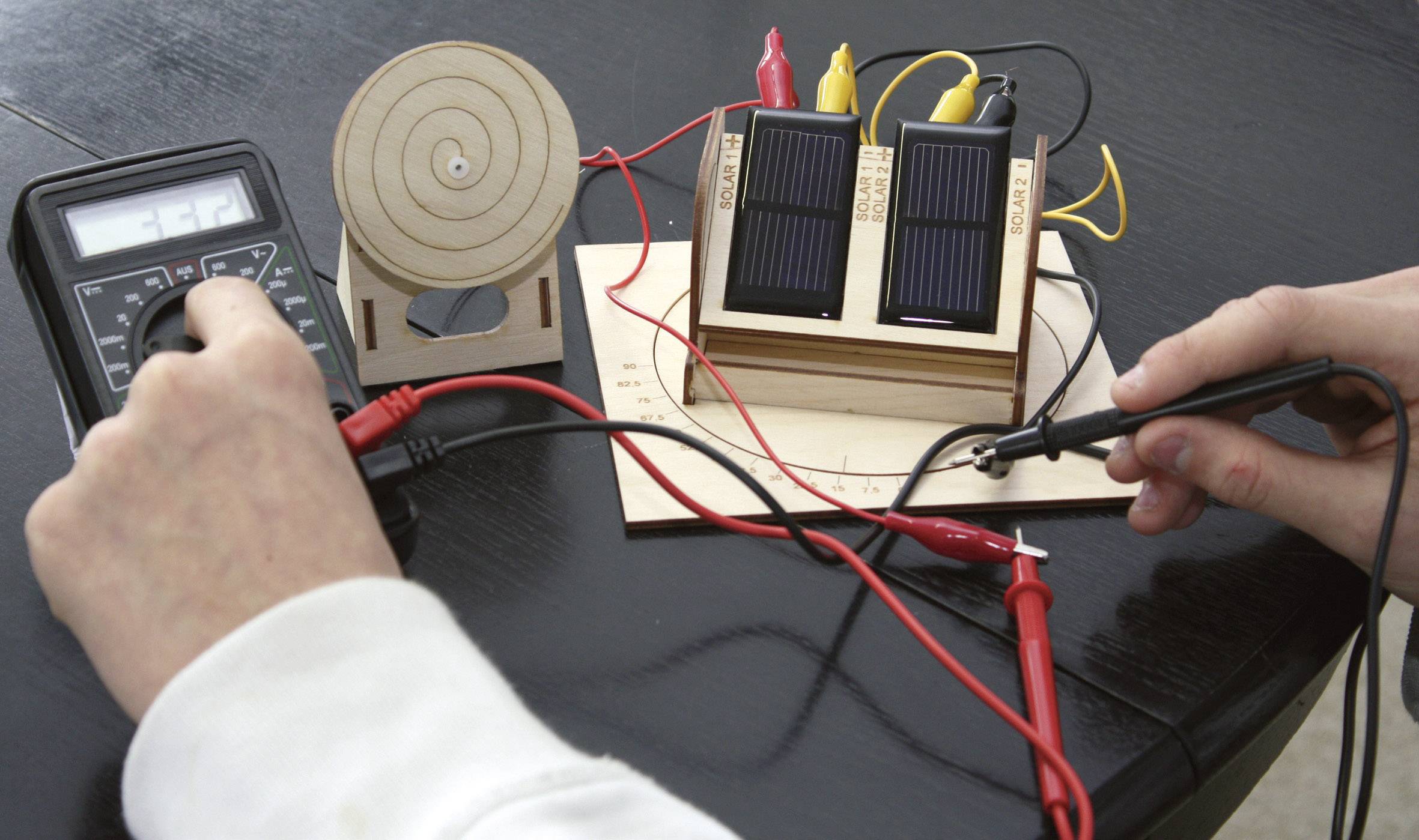 Une main tient un multimètre connecté à une expérience sur un panneau solaire ; il affiche la tension pendant que les modules solaires génèrent de l'énergie électrique.