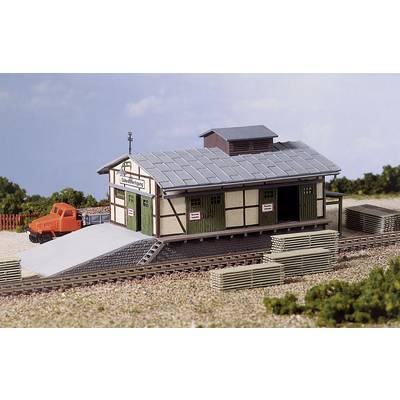 Un modèle d'une petite gare de marchandises avec un véhicule de levage orange à côté. Des arbres et un ciel bleu sont visibles en arrière-plan.