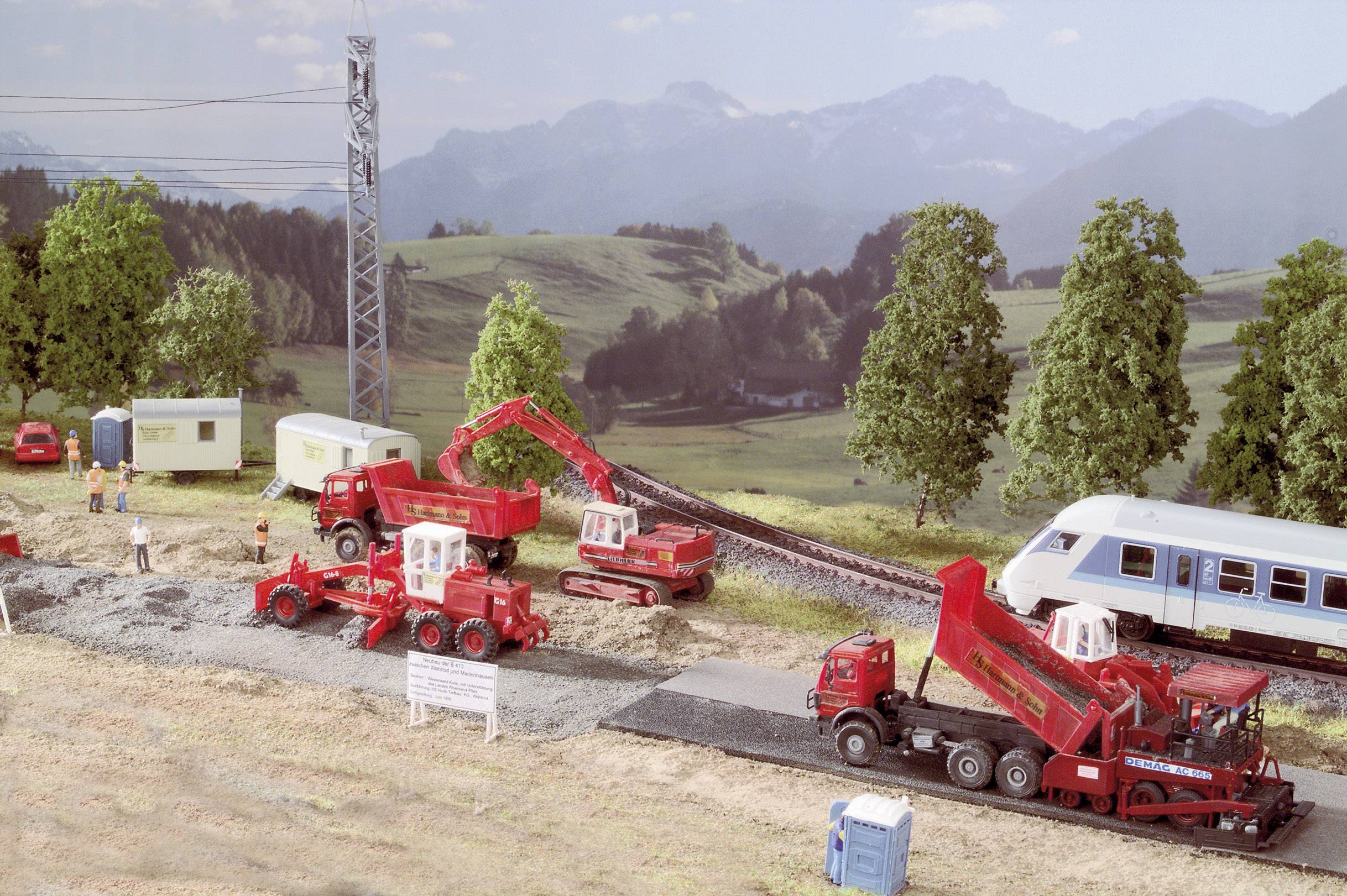 Un paysage miniature avec un chantier : Deux pelles mécaniques jouets travaillent sur une voie ferrée, tandis qu'une voiture de voyageurs est stationnée sur les rails.