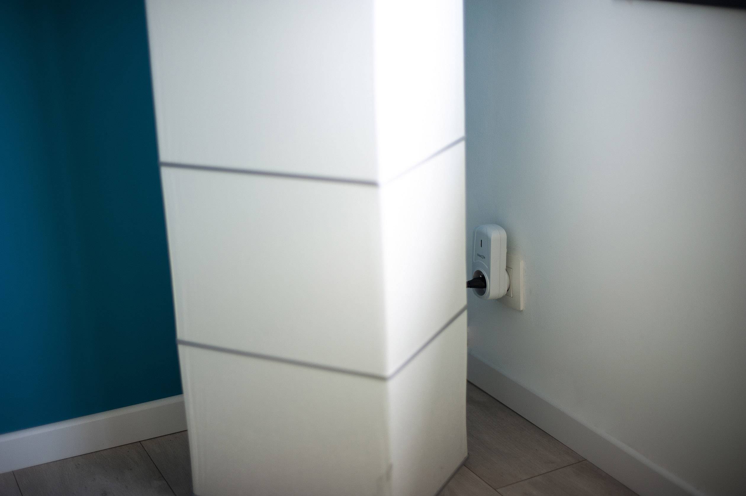 Un purificateur d'air dans un coin d'une pièce, près d'une prise électrique. Le mur est peint en bleu. Le sol est carrelé.