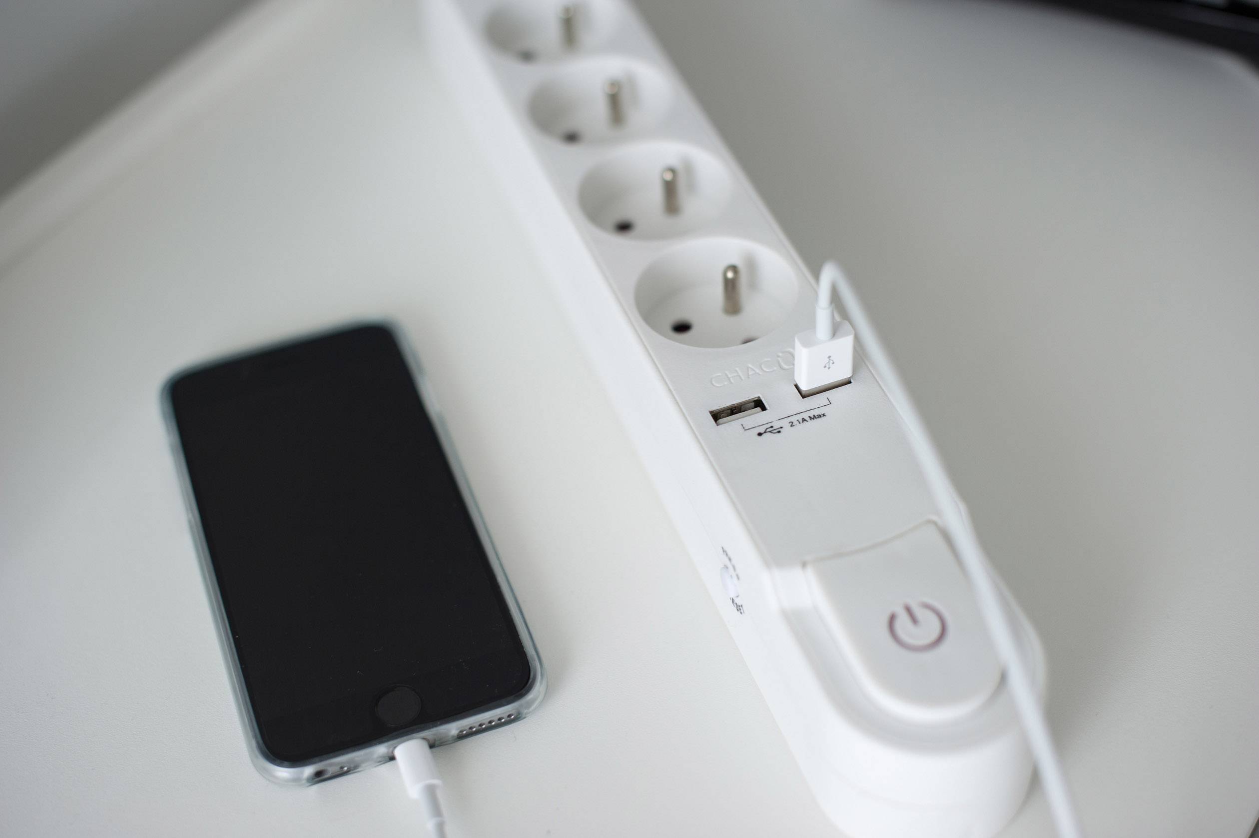 Un smartphone est posé à côté d'une multiprise qui branche un câble USB. La multiprise dispose de plusieurs prises et d'un grand interrupteur marche/arrêt.