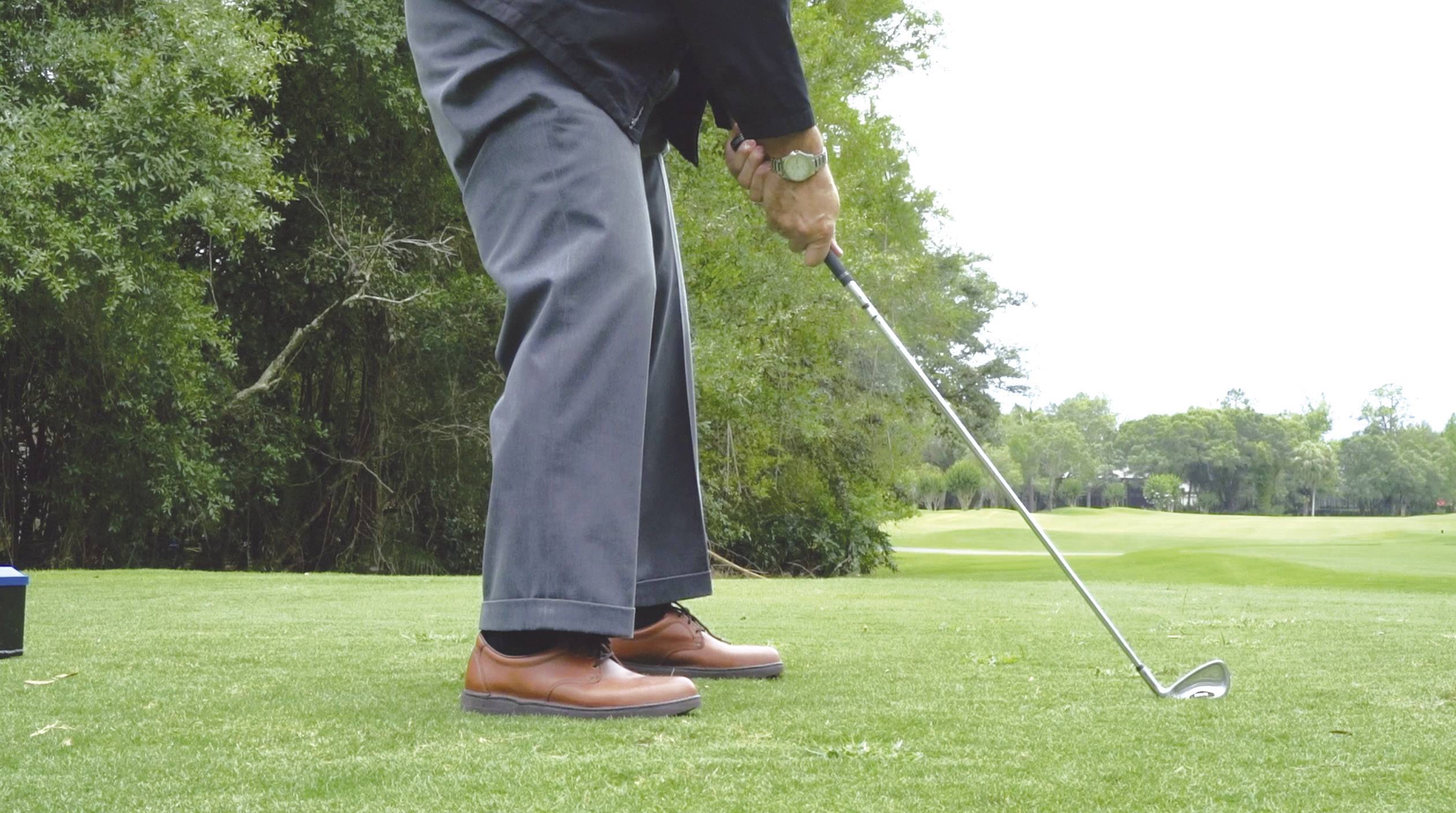 Une personne frappe une balle de golf sur un terrain de golf avec un club de golf. En arrière-plan, on peut voir de l'herbe et des arbres.